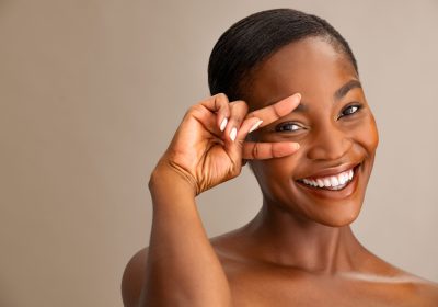 Portrait of smiling mature woman with perfect skin showing victory sign near eye on brown background. Closeup face of african woman with bare shoulder showing successful results after anti-aging wrinkle treatment. Crow's feet, eyebag and dark circles under the eyes concept.
